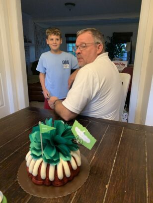 Man in white golf shirt looks over his shoulder to see a beautiful birthday cake with a green ribbon on it behind him. A boy with short light brown hair and a light blue shirt is facing him and the cake while smirking.