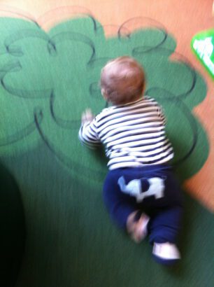 picture of a 6-8 month old baby crawling across a playroom floor with a green tree painted on it.  Baby is wearing navy blue and white striped long sleeved shirt and Navy pants with a light blue elephant on the butt area.