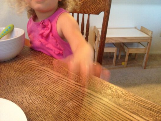 image of child with pink sleeveless shirt and ruffles rapping her hand on a wood table. Her hand appears to be in motion.