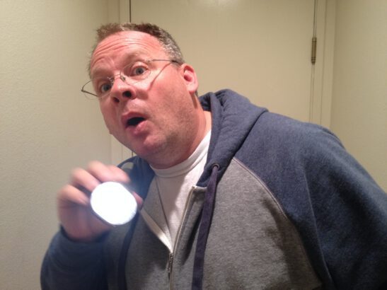 man with glasses and a gray and blue sweatshirt holds a flashlight to get someone's attention.  His facial expression shows he is looking toward the person, hoping to catch their gaze.