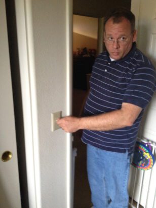 caucasian man with glasses gives a side-eye as he stands at at doorway flicking a wall light switch. He is wearing a navy blue striped golf shirt and jeans.