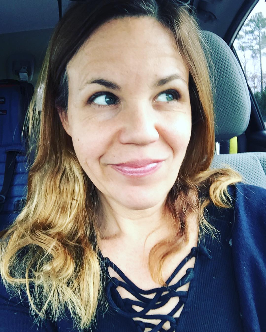 Jen sits in her car with a slight smirk on her face, looking upward thinking. She has golden brown hair with golden with loose waves around her shoulders. She is wearing a dark blue shirt with a criss cross thread design that rests loosely around the neckline.