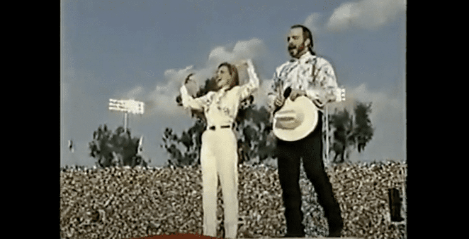Marlee Matlin performs the national anthem in ASL. She is dressed in a cream colored shirt and pants with a belt and has her arms raised in fists bent at the elbow. Garth Brooks sings next to her holding his cream colored cowboy hat and has a dark beard, light shirt and black pants. A large superbowl crowd is behind them.
