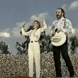 Marlee Matlin performs the national anthem in ASL. She is dressed in a cream colored shirt and pants with a belt and has her arms raised in fists bent at the elbow. Garth Brooks sings next to her holding his cream colored cowboy hat and has a dark beard, light shirt and black pants. A large superbowl crowd is behind them.
