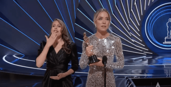 Sian Heder speaks into a microphone holding academy award trophy. She is wearing a shiny silver dress and blonde hair loosely tied back. A 2nd woman wearing a black dress and light brown loose curls in her hair signs thank you.