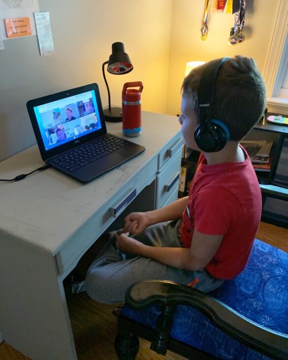 a young boy in a red shirt wears headphones and faces a laptop computer screen on his desk which shows other students in a zoom room.