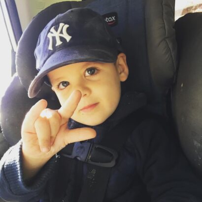 Caucasian toddler kid wearing dark jacket and NY Yankee baseball cap signs ASL I Love You while looking at the camera.