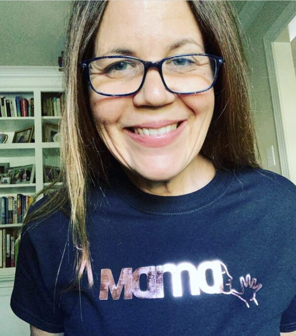 Caucasian woman with light brown hair and glasses leans toward and smiles at the camera. She is wearing a navy blue t shirt with a shiny pink MAMA decal in English and the American Sign Language sign emerging from the last letter A.