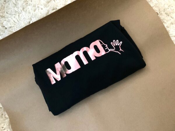 black shirt with pink foil decal spelling MAMA with the ASL sign emerging from the last letter A. shirt is folded into a rectangle and has been laid in the middle of brown paper on top of an off white shag rug.
