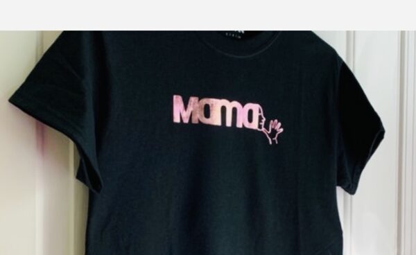 Black t shirt with a bright pink foil decal across the chest spells out MAMA with the ASL sign emerging from the last letter A. The shirt is hanging on an off white door.