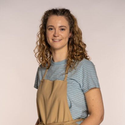 Tasha GBBO first deaf baker - a white woman with wavy sandy brown shoulder length hair smiles at the camera against a neutral backdrop. Tasha wears a light blue t-shirt and a tan apron. 