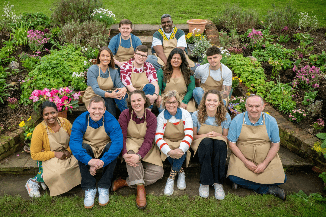 Does the GBBO Deaf Baker Go home - 12 GBBO bakers sitting on steps in a garden looking up at the camera and smiling. All are wearing tan aprons.