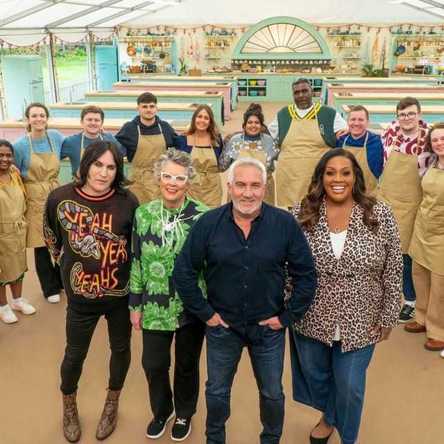 Great British bakeoff cast. 12 bakers in tan aprons stand in a semi circle behind 4 people smiling and looking up at the camera. A man with gray hair in front has his hands in his jeans pockets.