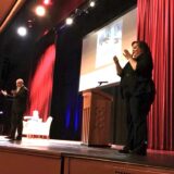 female ASL interpreter with black curly hair and black clothing stands on stage interpreting for a male speaker to her right.