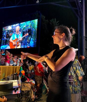 A female ASL interpreter interprets a musical performance as a screen behind her shows a male singing and playing a guitar.