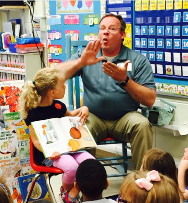 A Deaf Interpreter signs a story in ASL to a class of children as a little girl holds a book looking at him