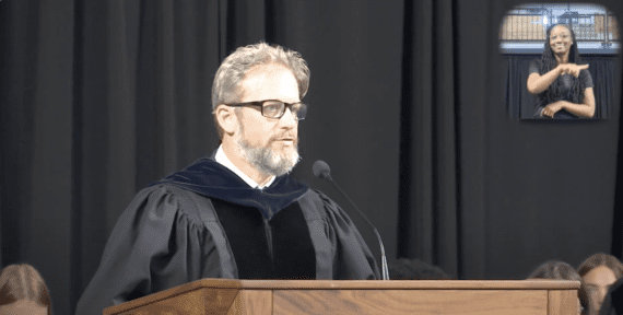 A bearded man with glasses wearing a black robe speaks into a microphone as he stands behind a podium in front of a black curtain. An ASL interpreter is shown in box in the upper right hand part of the screen.