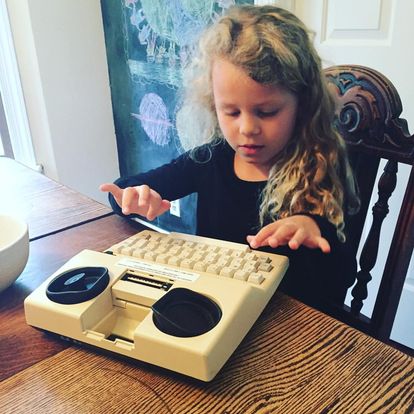 a young blonde girl with long curly hair sits at a table typing on an old TTY.
