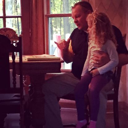 a Young KODA sits on her dad's lap at a table while he signs to her in ASL.