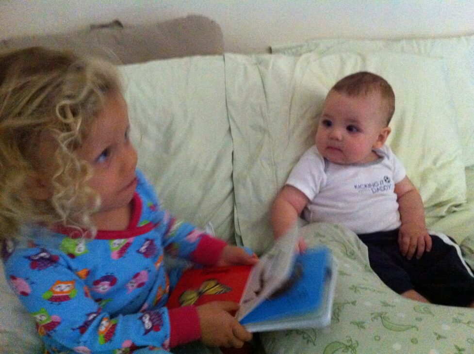 Young child reading to a younger sibling while sitting together on a couch